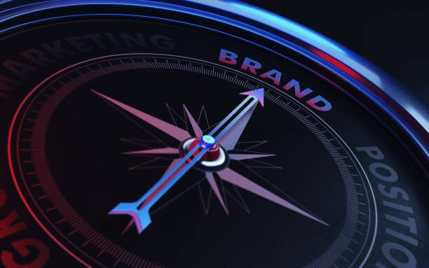 Arrow of a compass is pointing brand text on the compass. Arrow, brand text and the frame of compass are metallic blue in color. Red light illuminating compass is creating a sense of tension. Black backgound. Horizontal composition with copy space. Brand concept.