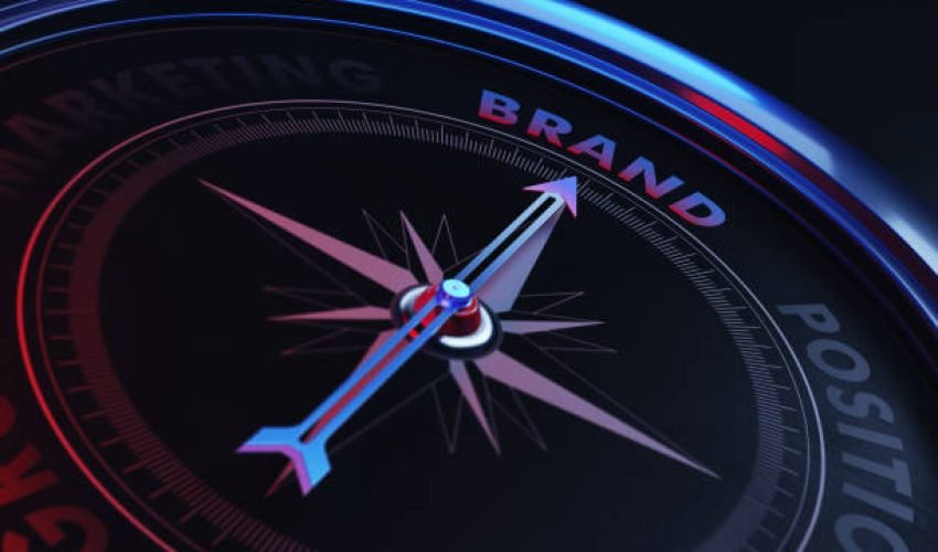 Arrow of a compass is pointing brand text on the compass. Arrow, brand text and the frame of compass are metallic blue in color. Red light illuminating compass is creating a sense of tension. Black backgound. Horizontal composition with copy space. Brand concept.